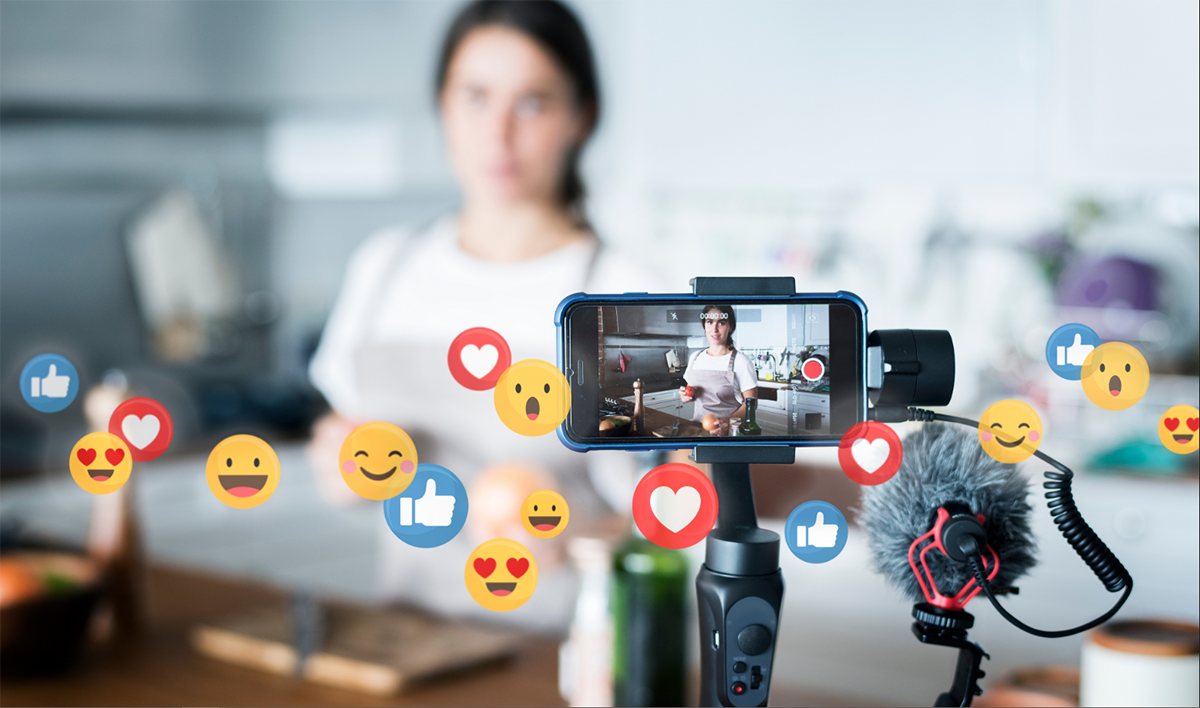 A woman in a white top and light brown apron is live streaming cooking in a kitchen.